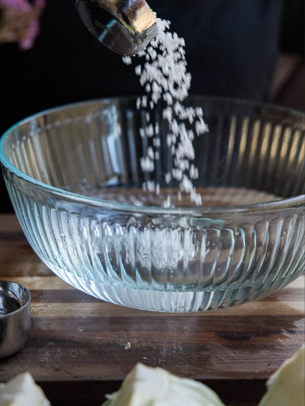 Salt being poured into a clear ribbed glass bowl on a wooden surface, with a metal measuring cup nearby.